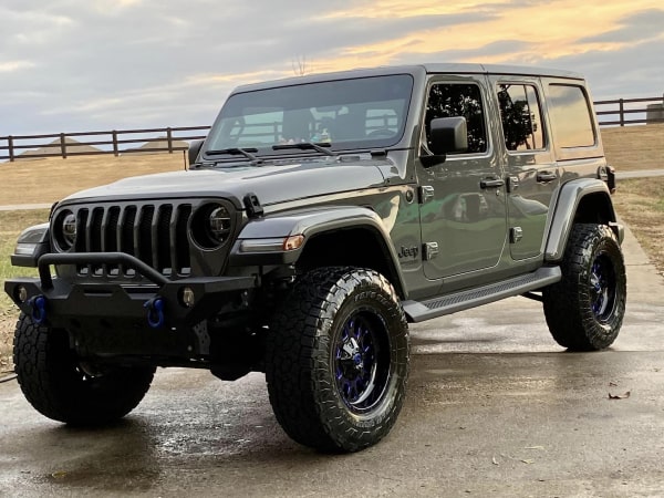 Modified Jeep Wrangler parked on a concrete driveway, featuring off-road tires, custom front bumper, and a clear sky background.
